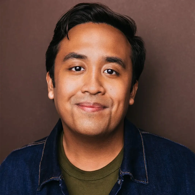 Gabriel Argate, a man with medium skin tone and dark hair, smiles gently at the camera. He is wearing a dark denim jacket over a green shirt, standing in front of a plain brown background with even lighting highlighting his facial features.