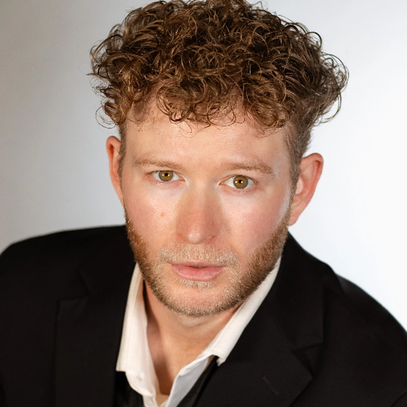 A close-up portrait of Ethan Hardy Benson, a man with light-brown, tightly curled hair and hazel eyes, wearing a black blazer over a white collared shirt. He has a light beard and is looking directly at the camera with a neutral expression against a simple, bright, light-gray background.