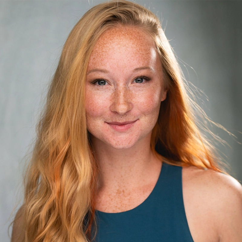 A young woman with long wavy strawberry-blonde hair and freckles smiles gently at the camera. She wears a sleeveless teal top and is standing in front of a softly blurred gray background. Soft lighting highlights her natural features.