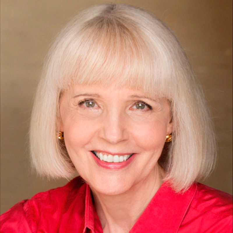 A smiling woman with straight, chin-length blonde hair and bangs wears a bright pink collared shirt. She has fair skin, light-colored eyes, and small, gold, square-shaped earrings. The background is plain and softly lit in warm beige tones.