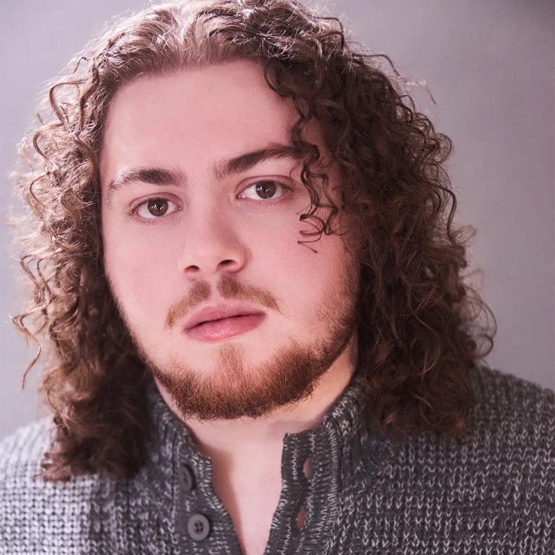 Quinn Dembecki, a person with long, curly brown hair and a trimmed beard, looks at the camera with a neutral expression. They wear a textured grey buttoned sweater and are posed against a softly lit, neutral background.