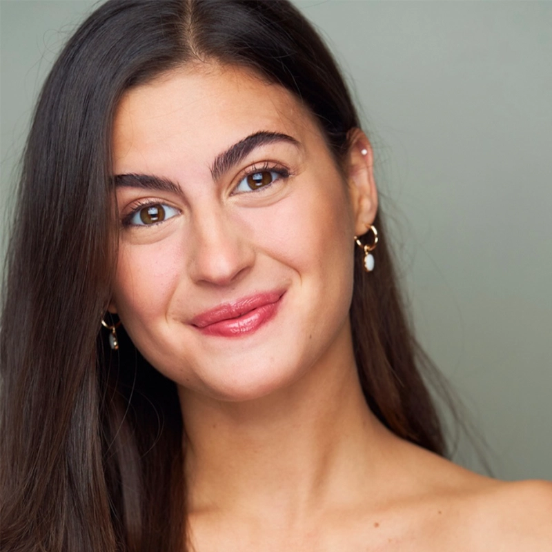 A young woman with long, straight dark brown hair smiles gently. She has clear skin, arched eyebrows, and wears subtle makeup with glossy pink lipstick—a portrait styled by Ethan Hardy Benson. She wears small gold hoop earrings against a light gray background.