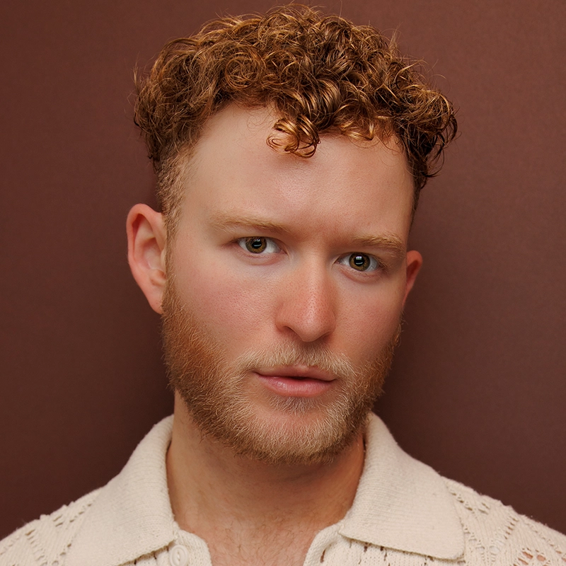 Ethan Hardy Benson, a young man with fair skin, short curly reddish-brown hair, and a trimmed beard, poses against a plain brown background. He wears a cream-colored textured collared shirt and looks directly at the camera with a neutral expression.