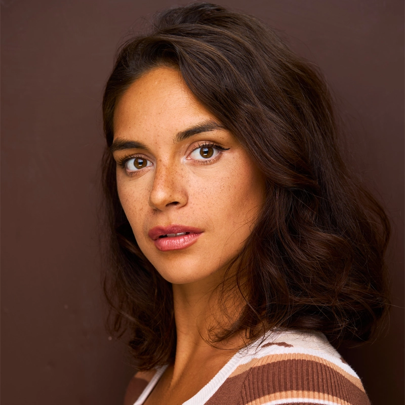 A young woman, Jo Garcia-Reger, with medium skin tone and wavy dark brown hair looks directly at the camera. She has brown eyes, natural makeup, and wears a brown-and-white striped top against a plain brown background.