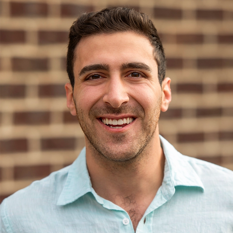 A young man with short dark hair and trimmed facial hair smiles warmly at the camera. He wears a light blue button-up shirt and stands in front of a blurred brick wall background. The lighting is natural and even.