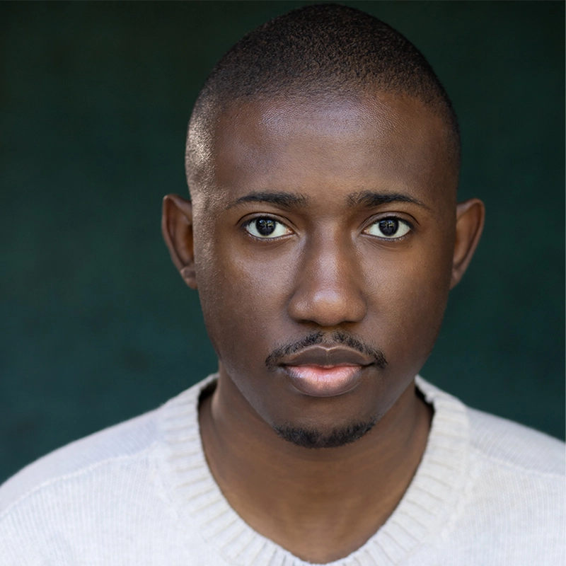 A young Black man with short hair and a trimmed mustache and goatee looks directly at the camera. He is wearing a light beige sweater and stands against a dark green, softly blurred background. The lighting is even, highlighting his facial features.