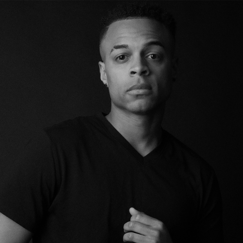 A young man with short curly hair, wearing a black V-neck shirt, stands against a dark background. He looks directly at the camera with a neutral expression. His left hand is partially raised near his chest. The lighting is soft and highlights his facial features.