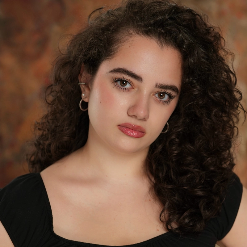 Lauren A. Marchand, a young woman with long, curly dark hair and fair skin, looks at the camera with a neutral expression. She wears a black scoop-neck top and hoop earrings, set against a softly blurred background in warm brown and orange tones.