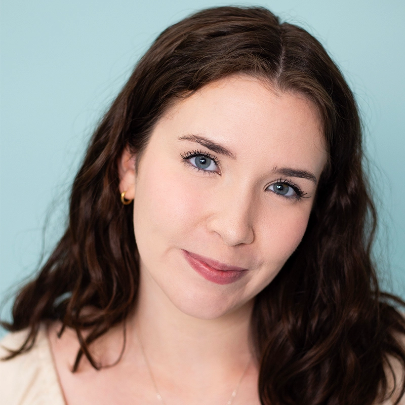 A young woman with long, wavy brown hair and blue eyes smiles softly. She wears a light top and a small gold hoop earring. The background is a solid pale blue, reflecting the calm, gentle style favored by Ethan Hardy Benson. Her expression is warm and relaxed.