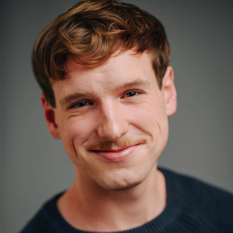 Ethan Hardy Benson, a young man with light skin, short wavy brown hair, and a light mustache, smiles softly. He wears a dark blue sweater against a smooth, neutral gray background. Soft, even lighting keeps the focus on his face.