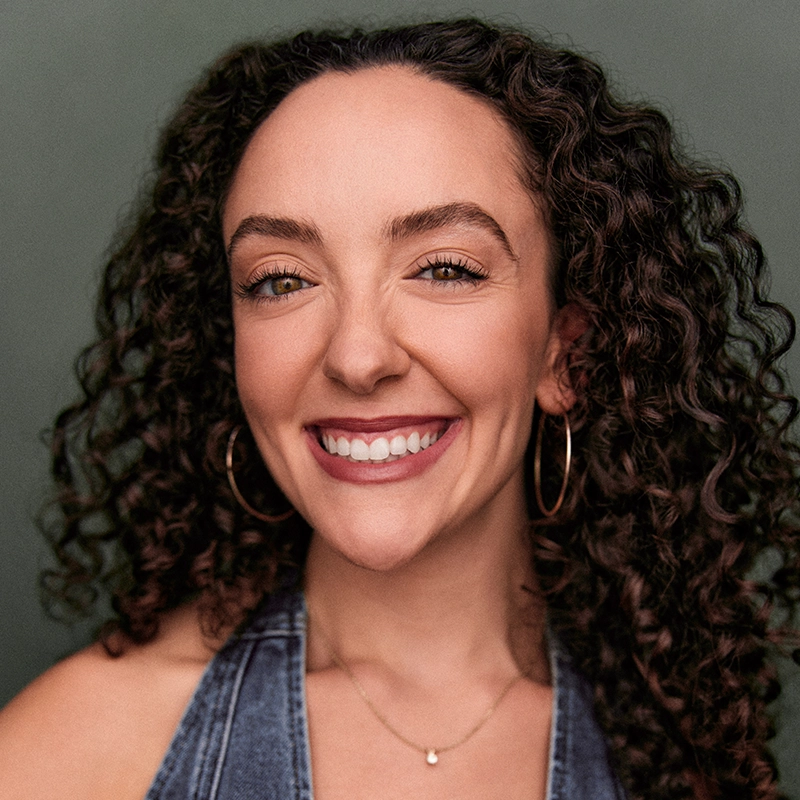 A woman with curly brown hair smiles broadly, showing her teeth. She wears large hoop earrings and a blue sleeveless top. The lighting highlights her cheerful expression in a muted green setting reminiscent of an Ethan Hardy Benson portrait.