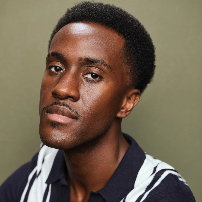 Zion Middleton, a man with short, neatly trimmed hair and a faint mustache, looks at the camera with a neutral expression. He is wearing a navy blue collared shirt with white stripes against a plain olive green background.