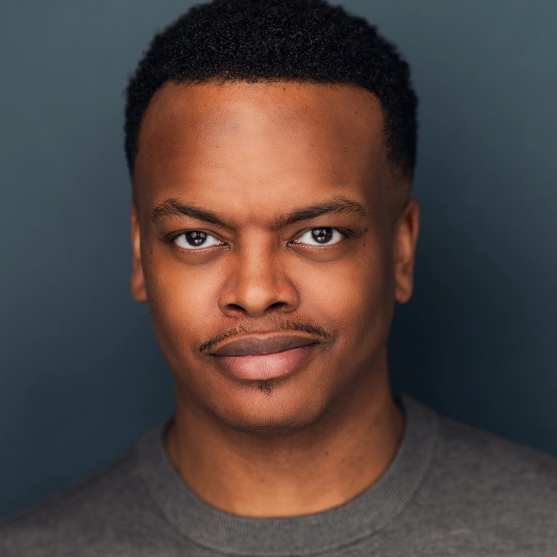 Michael Nero, a man with short curly hair and a thin mustache, wears a gray crewneck shirt as he faces forward, smiling slightly. The soft, even lighting highlights his features against a smooth, dark blue-gray background.
