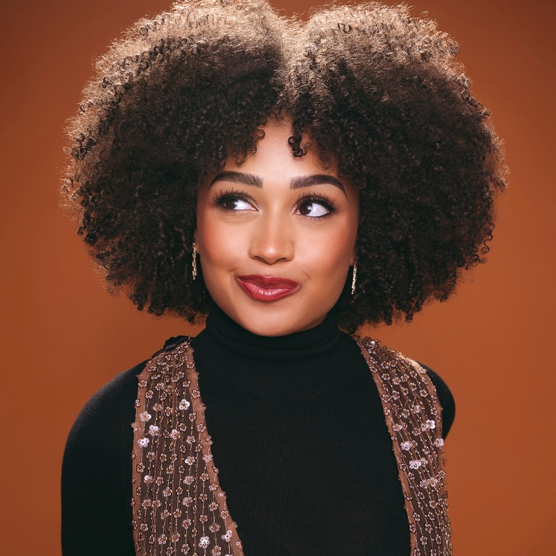 Jasmine Amy Rogers, with voluminous curly hair, looks slightly to the side, smiling softly. She wears a black turtleneck and a sleeveless, sequin-embellished top against a warm orange background, her bold red lipstick completing the look.