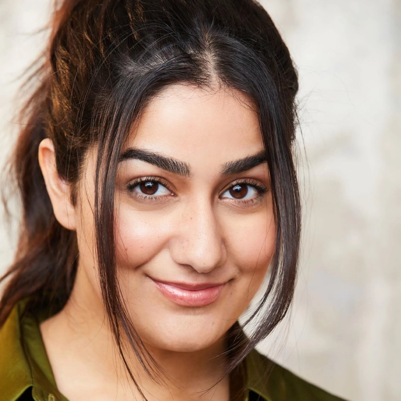 Soleil Singh, a woman with long dark hair tied back and loose strands framing her face, smiles gently. She has full eyebrows, brown eyes, smooth skin, and wears a green collared shirt against a softly blurred neutral background.