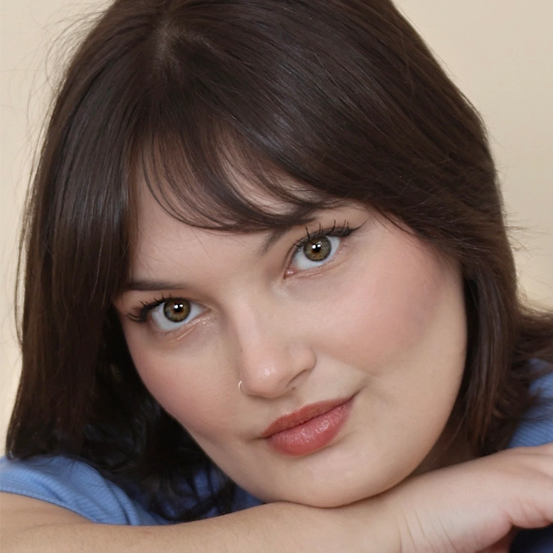 A woman with short brown hair and hazel eyes gazes directly at the camera, resting her chin on her crossed arms. She has natural makeup, a small nose ring, and wears a soft blue shirt—captured by photographer Ethan Hardy Benson.
