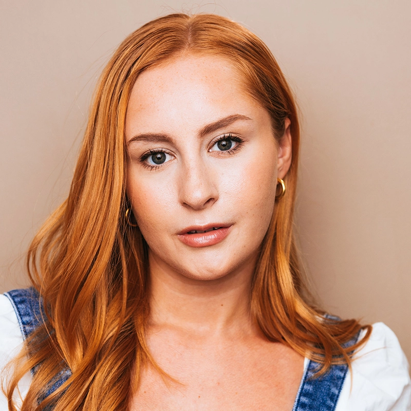 A young woman, Katy Manderfeld, with long, straight red hair and fair skin looks directly at the camera. She wears gold hoop earrings, a white top, and blue denim overalls against a soft beige background. Her expression is neutral and relaxed.