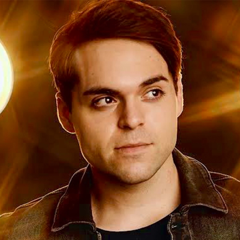 A young man with fair skin and short brown hair looks slightly to his left. He wears a dark jacket over a black shirt. The background glows with dramatic amber lights, casting a calm, thoughtful mood reminiscent of Chelsea Udoye’s evocative portraits.