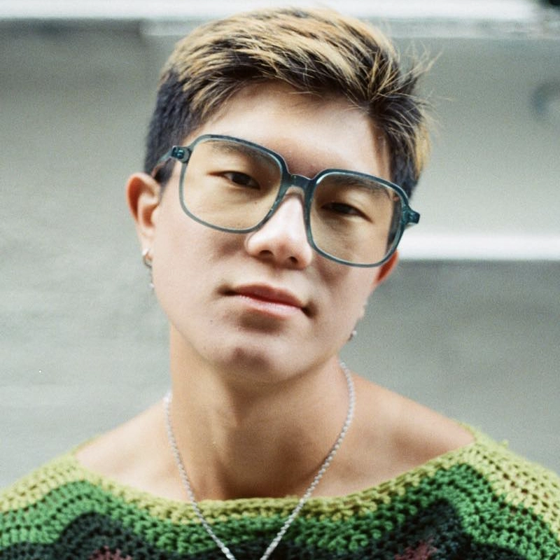 Aeden Alvarez, a young person with short, dark hair featuring blond highlights, wears large translucent teal glasses and a green crocheted sweater. Silver earrings and a necklace accent their look as they gaze at the camera with a neutral expression.