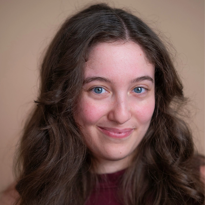 A young woman with long wavy brown hair and blue eyes smiles gently at the camera. She wears a sleeveless burgundy top and is posed against a plain, light beige background. Her expression is warm and inviting, capturing the approachable charm often seen in Amanda Ribnick’s portraits.