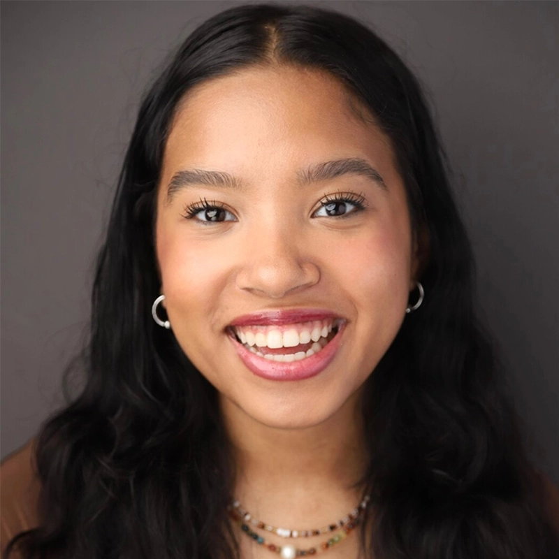 A young woman with long, wavy dark hair smiles brightly at the camera. She wears hoop earrings and layered bead necklaces. Her skin is smooth and she has natural makeup. The background is a plain, neutral gray, highlighting Katy Manderfeld’s vibrant style.