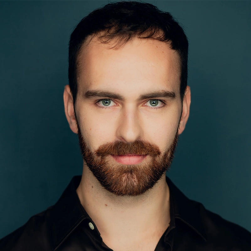 A man with short dark hair, a full reddish-brown beard, and a mustache is facing forward, smiling slightly. He has light eyes and wears a dark collared shirt. The background is a solid dark teal color, reminiscent of Katy Manderfeld’s style.