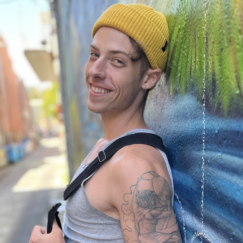 Mitch Bowers, a young man in a yellow beanie, gray tank top, and tattoo on his upper arm, smiles while leaning against a colorful painted wall in an outdoor urban alley, with blurred buildings and greenery in the background.