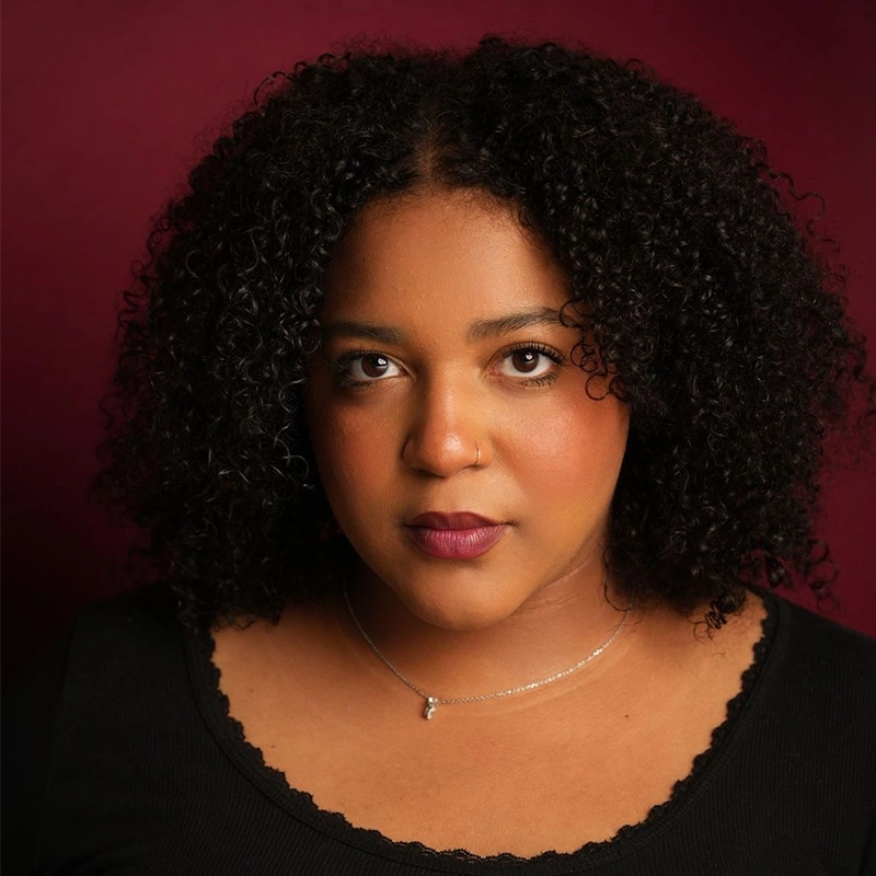 Hannah Adams, a woman with medium-dark skin and curly black hair, looks at the camera with a neutral expression. She wears a black top with scalloped edges and a delicate silver necklace. The background is a deep burgundy color, while the lighting softly highlights her face.