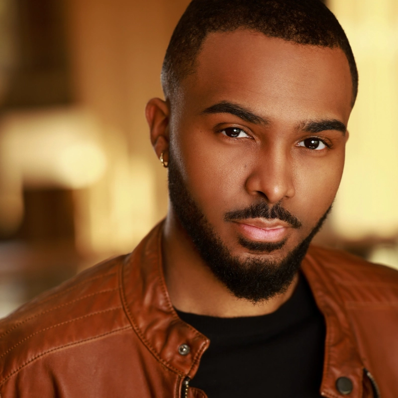 JaVaun Ryal Butler, a man with short hair and a neatly groomed beard, looks at the camera with a neutral expression. He is wearing a brown leather jacket over a black shirt, set against a softly blurred background in warm tones.