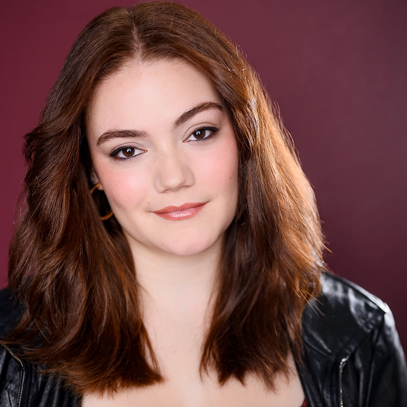 A young woman with fair skin and shoulder-length brown hair smiles softly. Hannah Adams wears a black leather jacket and hoop earrings. Her makeup is subtle, with pink blush and natural lip color. The background is a deep maroon shade.