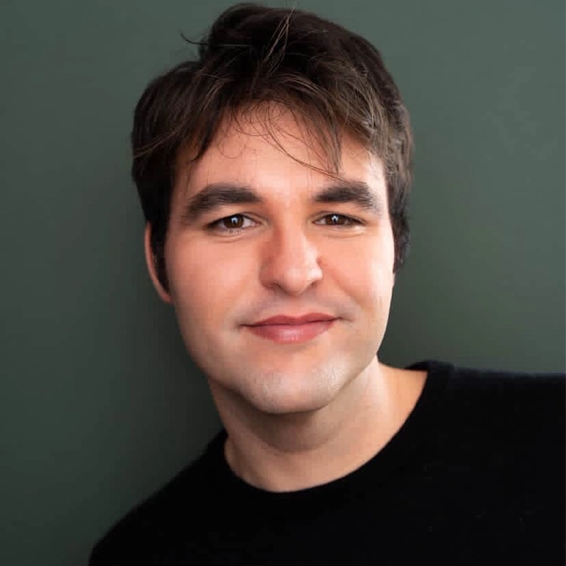 Julian Carli, a young man with fair skin, short brown hair, and brown eyes, smiles softly. He wears a black top and poses against a dark green background, his slightly tousled hair framing a relaxed, friendly expression.