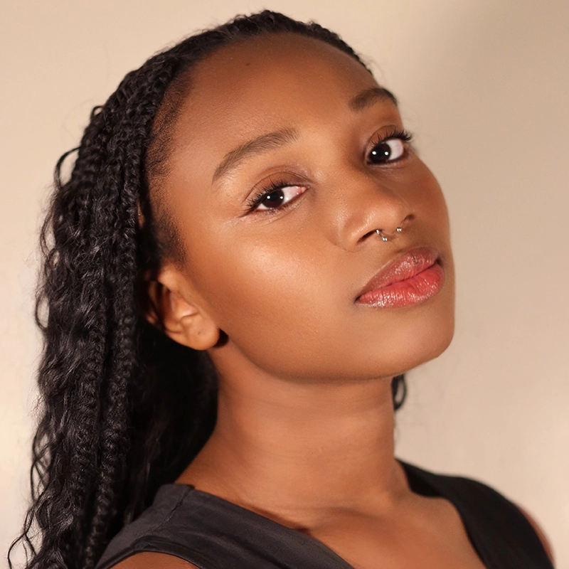 Chelsea Udoye, a young woman with long, braided black hair, poses against a neutral background. She has smooth brown skin, glossy red lips, natural makeup, a septum piercing, and exudes confidence in her black sleeveless top.