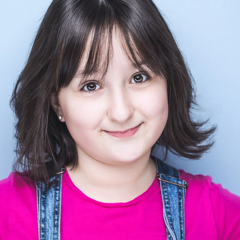 A young girl with straight, dark brown hair and bangs smiles gently at the camera. She wears a bright pink shirt under blue denim overalls. The background is light blue, giving the portrait a soft, Violet Winter charm as her hair is tucked behind one ear.