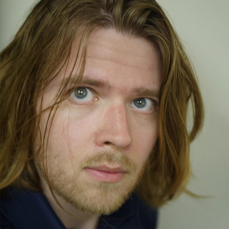 A person with long, light brown hair and blue eyes looks slightly upward toward the camera. They have light facial hair and wear a navy blue shirt. The background is a plain, light color, keeping the focus on their face—reminiscent of Amanda Ribnick's signature minimalist portrait style.