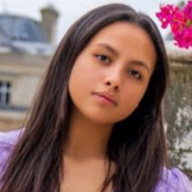 A young woman with long, straight brown hair and a calm expression looks slightly past the camera. Katy Manderfeld wears a lavender top and stands outdoors near a stone railing and bright pink flowers, with blurred buildings in the background.