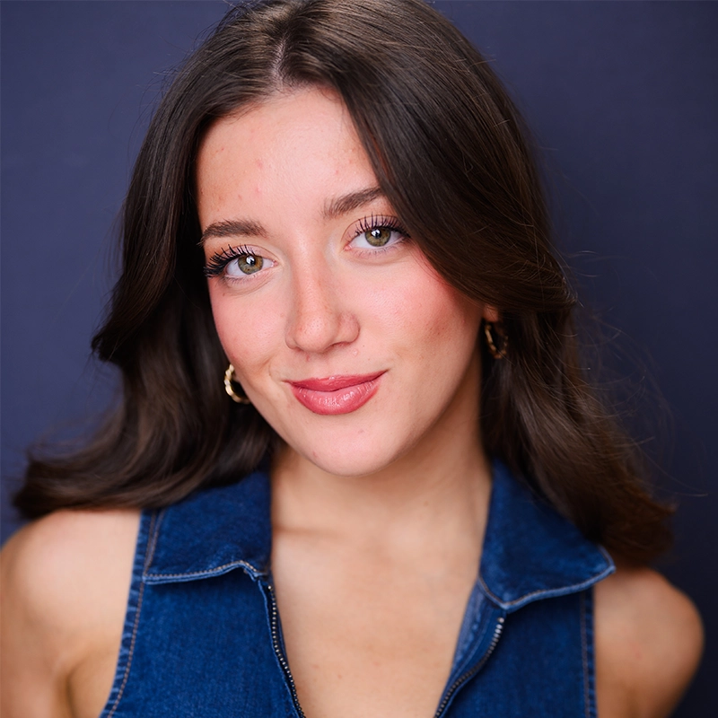 Hannah Adams, a young woman with long brown hair and light eyes, smiles softly. She wears a sleeveless blue denim top and gold hoop earrings. The background is dark blue, and her makeup is natural, with subtle pink lipstick and visible eyelashes.