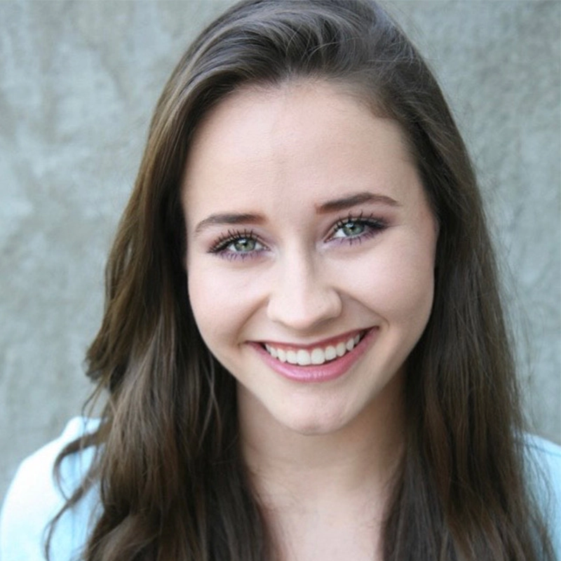 A young woman with long brown hair and blue eyes smiles warmly at the camera. She is wearing a light blue top, and the background is a plain, textured gray wall. Her makeup is natural, highlighting her eyes and bright smile—an inviting portrait of Amanda Ribnick.