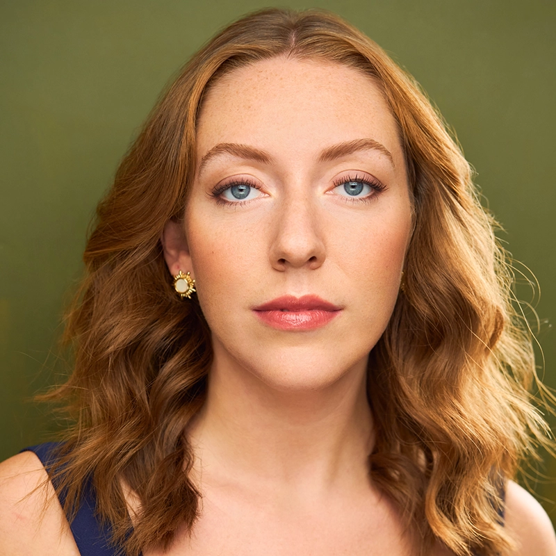 Katy Manderfeld, with wavy, shoulder-length auburn hair and blue eyes, faces the camera with a neutral expression. She wears gold flower-shaped earrings and a navy top against a plain, olive green background, softly and evenly lit.
