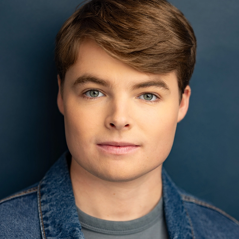 A young person, Katy Manderfeld, with short, light brown hair and blue eyes faces the camera against a dark blue background. They wear a denim jacket over a gray shirt and have a neutral, calm expression. Soft lighting highlights their features evenly.