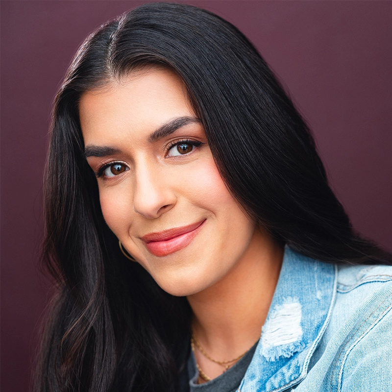 Chelsea Udoye, with long, straight dark hair, smiles gently at the camera. She wears natural makeup with light pink lipstick and a light blue denim jacket over a dark top, set against a solid dark maroon background.