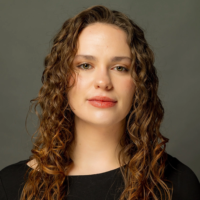 Hannah Adams, a woman with fair skin, wavy brown hair, and hazel eyes, faces the camera against a gray background. She wears a black top and has a neutral expression, with subtle makeup and slightly parted lips.