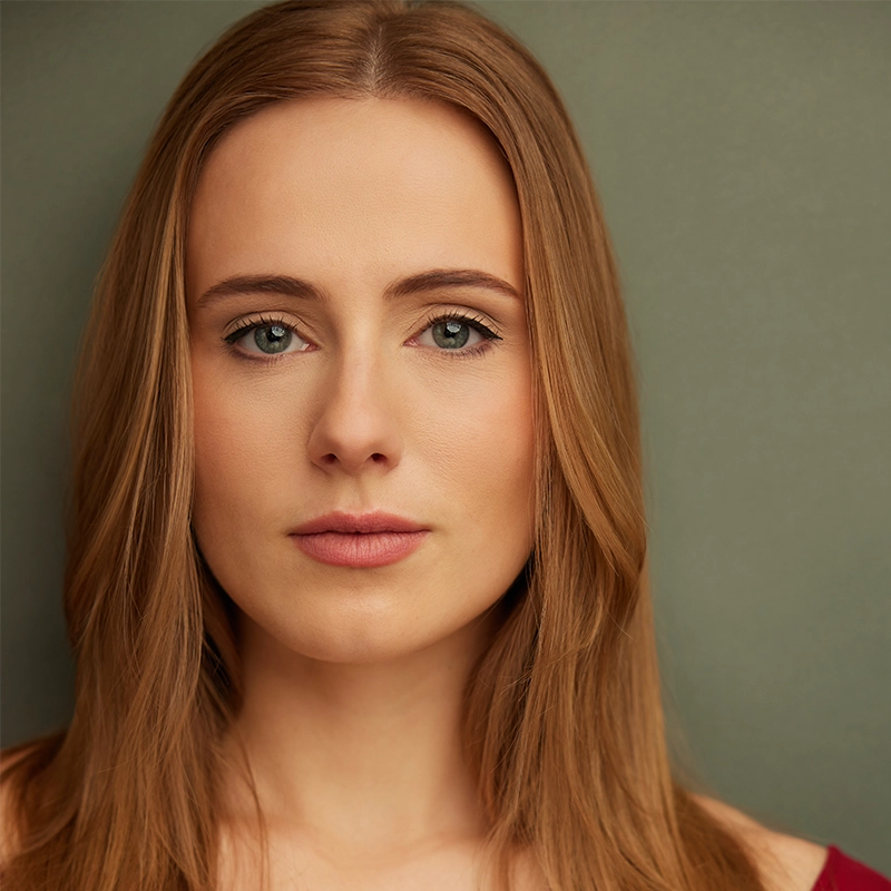 A woman with straight, light brown hair and fair skin looks directly at the camera. She has green eyes, natural makeup, and a neutral expression. The background is plain and muted green, and she is wearing a burgundy top. Amanda Ribnick’s poised presence is subtly highlighted in this understated portrait.