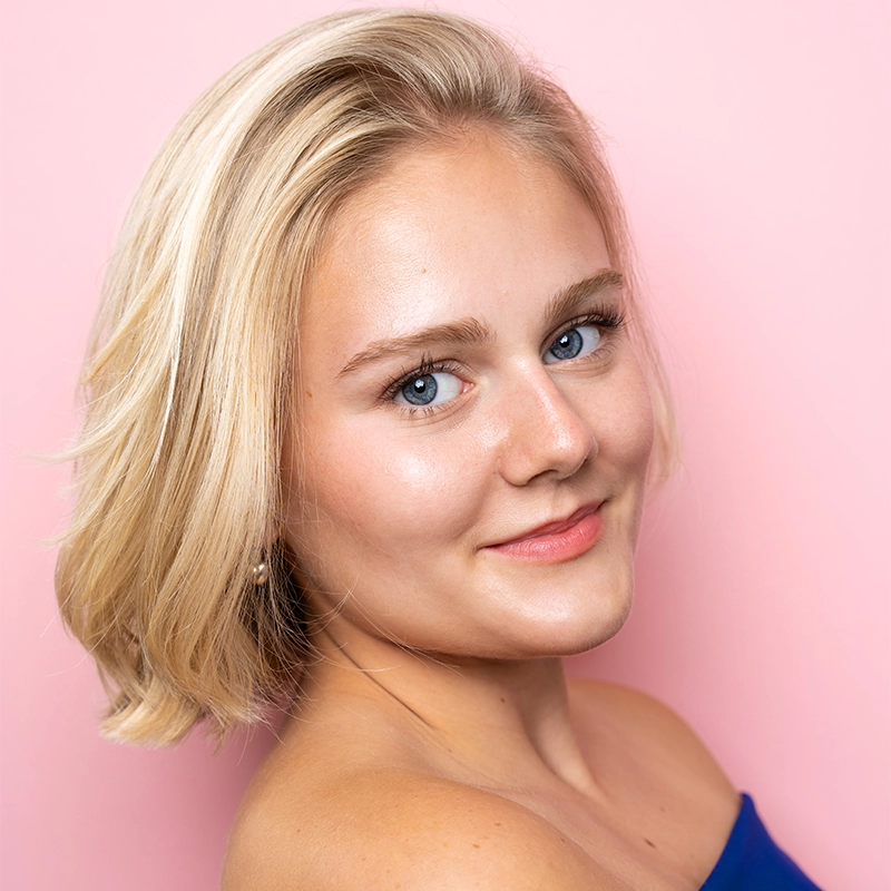 A young woman with short, blonde hair and blue eyes smiles softly at the camera. Wearing a blue off-the-shoulder top, Katy Manderfeld’s subtle, natural makeup stands out against the solid light pink background, giving the portrait a cheerful feel.