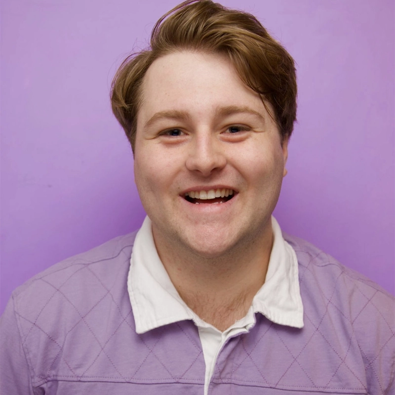 A young man with light skin and short, light brown hair smiles at the camera. Joseph Keegan is wearing a lavender quilted sweater over a white collared shirt. The background is solid light purple.