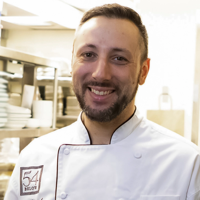 A smiling man with short brown hair and a beard, identified as James Klapak, wears a white chef’s coat with a “54 Below” patch. He stands in a bright kitchen with shelves of dishes and metal cookware visible in the background.