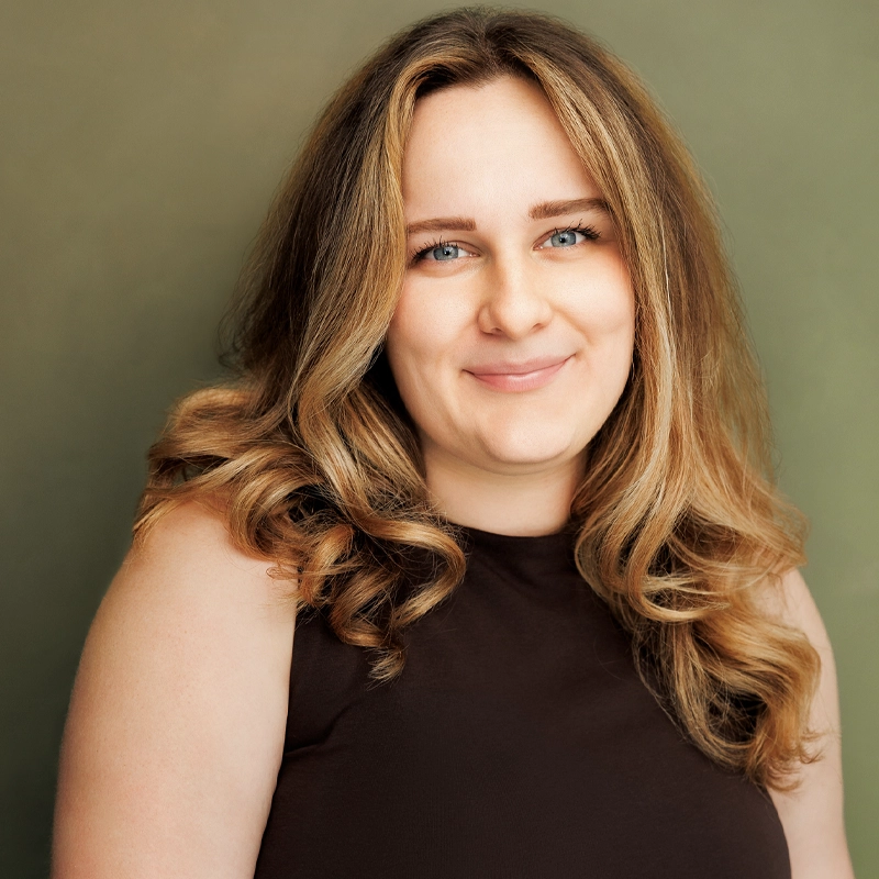 A woman with long, wavy blonde hair smiles at the camera. She wears a sleeveless dark brown top and is posed in front of a soft green background. The lighting is warm and even, highlighting Kamryn Koerner’s friendly expression.