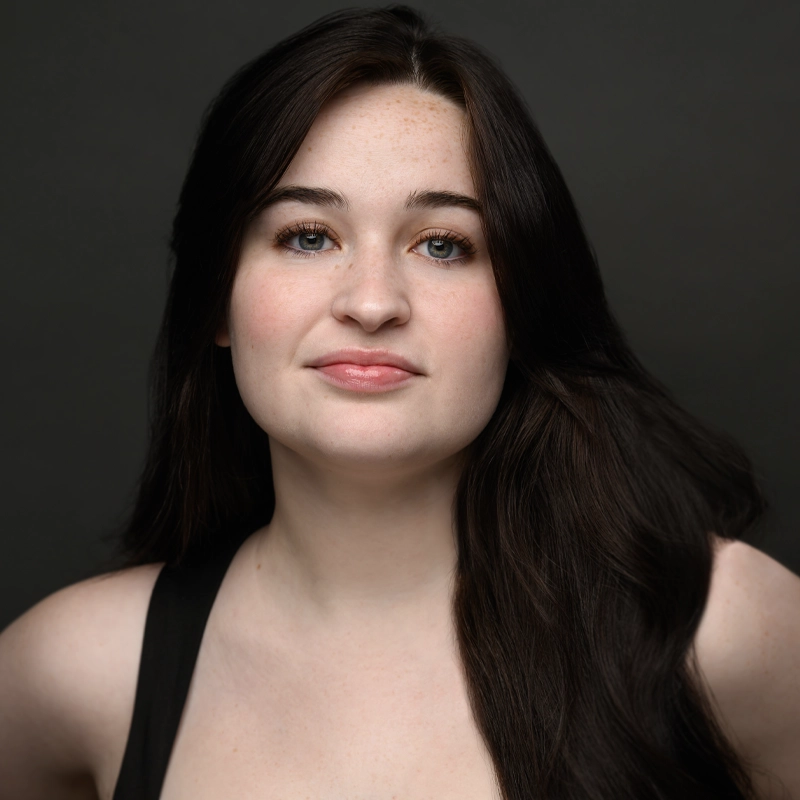 A young woman with long dark brown hair and fair skin poses against a dark background. With blue eyes, light freckles, and a subtle smile, Kylie Lavrenchik wears a sleeveless black top and looks confidently toward the camera.