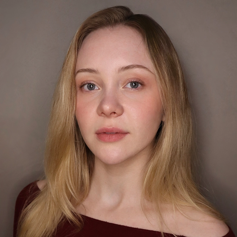 A young woman with long blonde hair and fair skin looks at the camera with a neutral expression. She wears a burgundy off-shoulder top, with minimal makeup. The background is plain and taupe. Soft lighting highlights her face evenly, drawing attention to her delicate features—capturing Hannah Adams in a poised and understated moment.