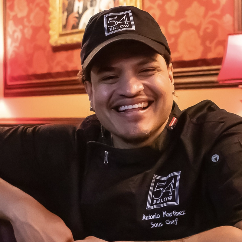 A smiling Antonio Martinez, wearing a black chef’s uniform and a hat embroidered with “54 Below,” sits in a warmly lit room with red patterned walls and framed art. His uniform displays his name and title: 