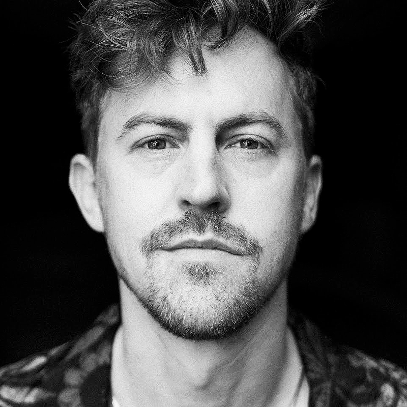 A black-and-white close-up portrait of a young man with wavy hair, light stubble, and mustache. He gazes directly at the camera with a neutral expression. The background is dark, making his face the main focus of the image.